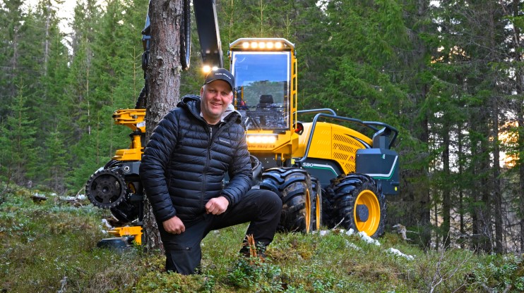 Machine operator Jahn Steinar kneels in front of his yellow forestry machine surrounded by trees and woodland.