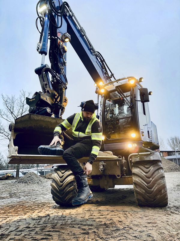 A man dressed in work gear sits in the bucket of his excavator.