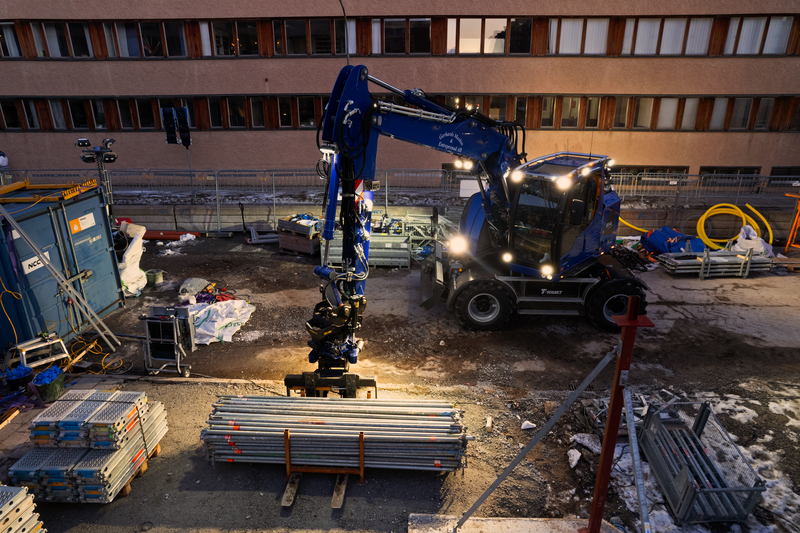 A blue excavator works on a construction site at night with its work lights lighting up the site.